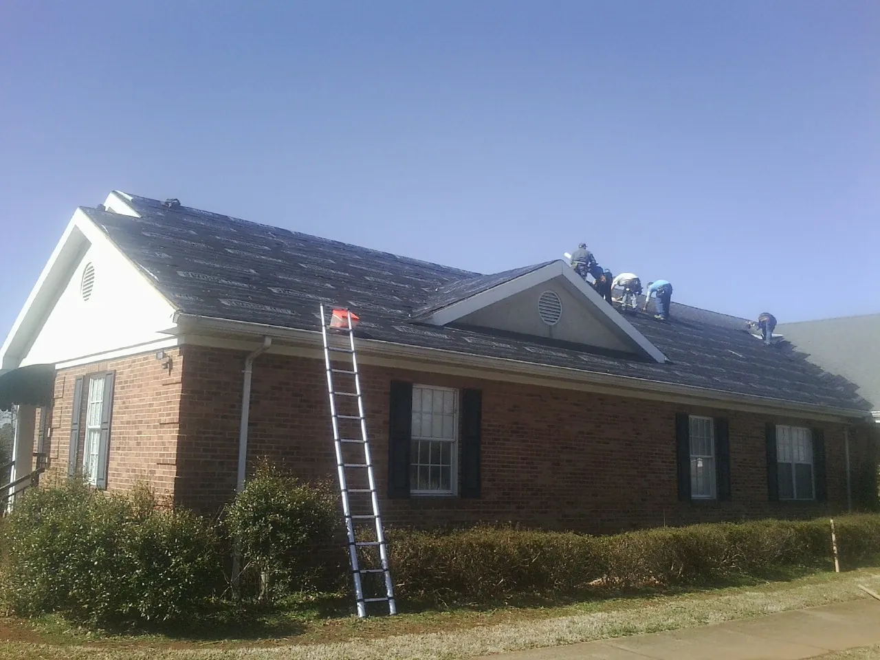 Crew tearing shingles off a brick ranch roof with a ladder set — Emergency Roof Repair work by our crew in Saint Johnsbury