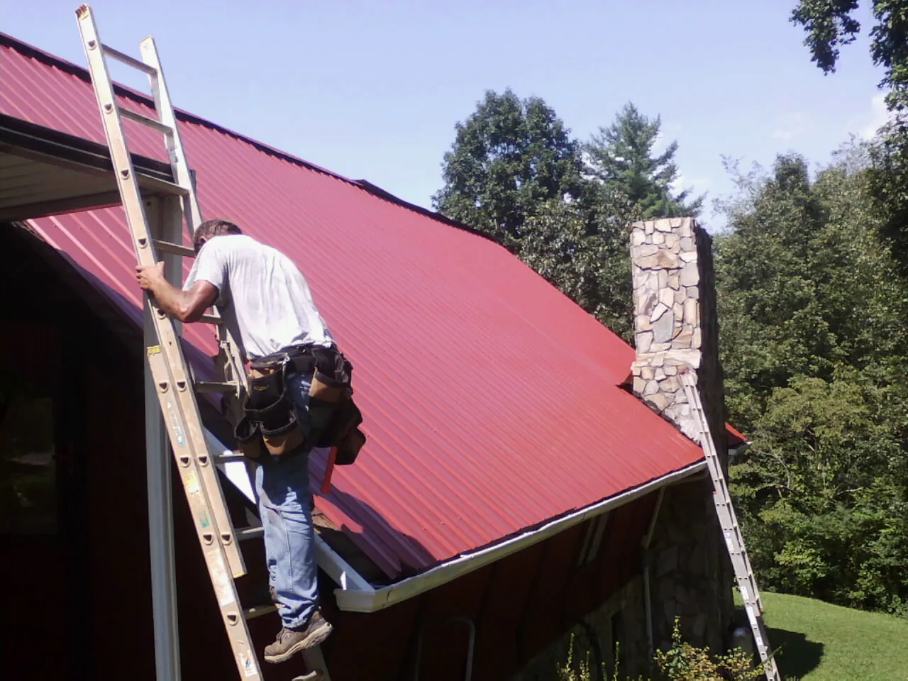 Roofer on a ladder beside a red metal roof and stone chimney — Soffit & Fascia Repair by Roofing Brothers in Saint Johnsbury